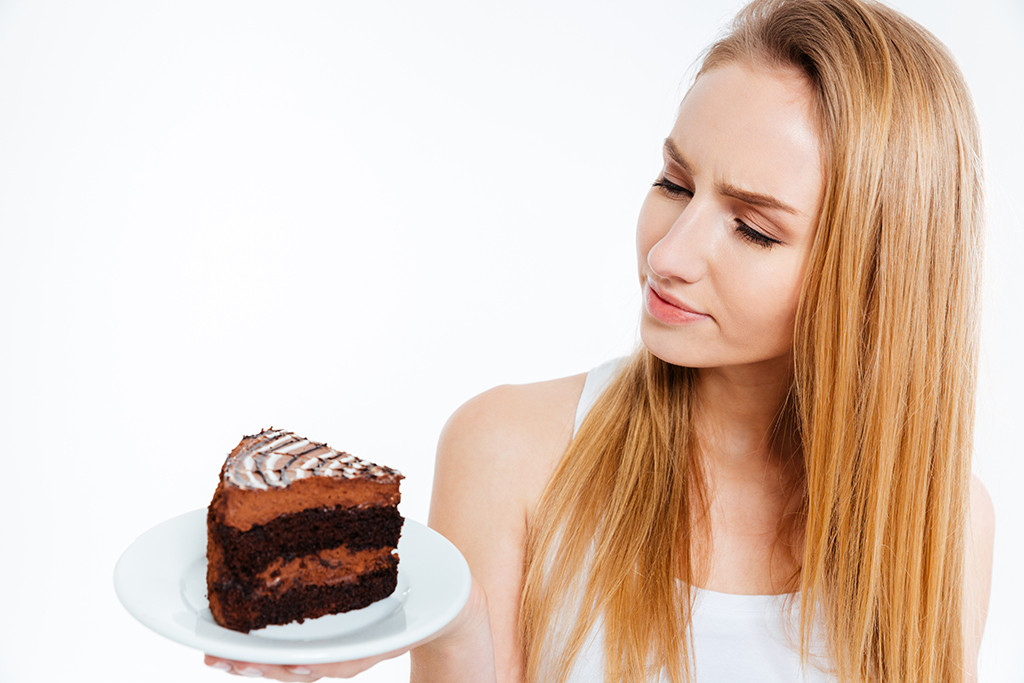 Pensive beautiful young woman looking at piece of chocolate cake over white background